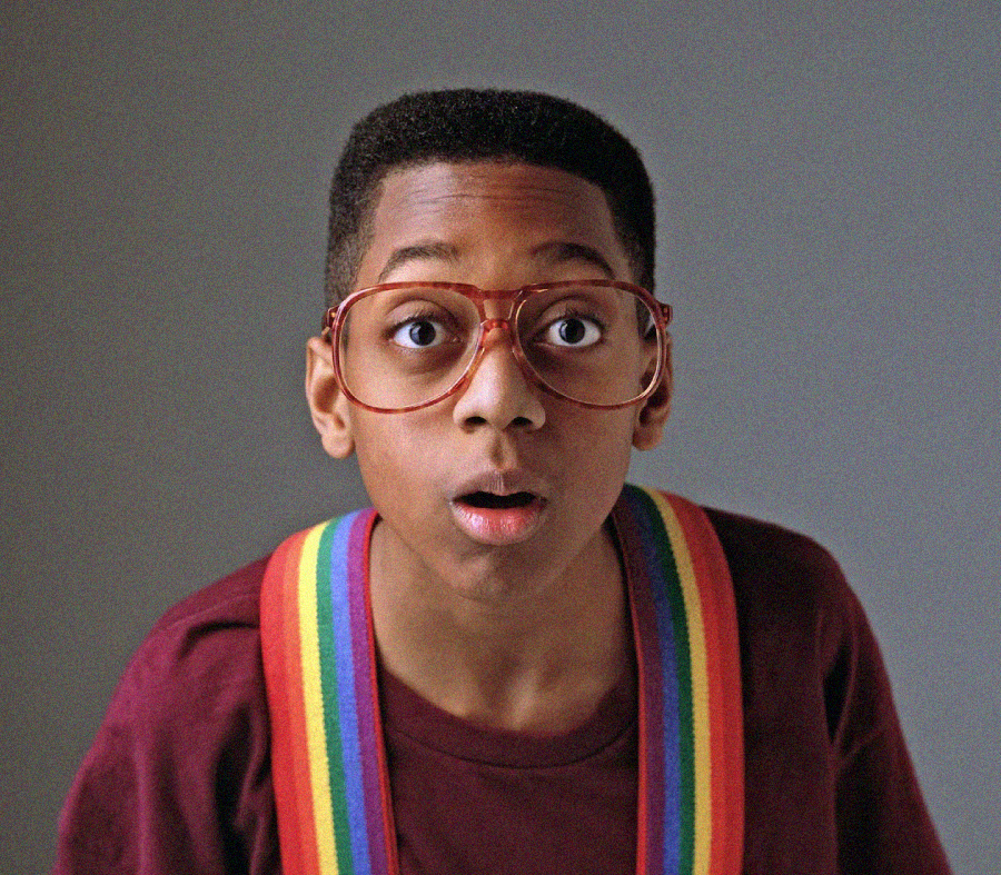 A young boy with a surprised expression, wearing oversized red glasses, a maroon shirt, and rainbow suspenders, stands against a plain gray background.