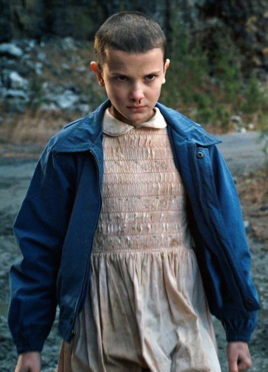 A young girl with a shaved head wears a blue jacket over a light pink dress, standing outdoors with a serious, intense expression. Trees and rocks are visible in the background.
