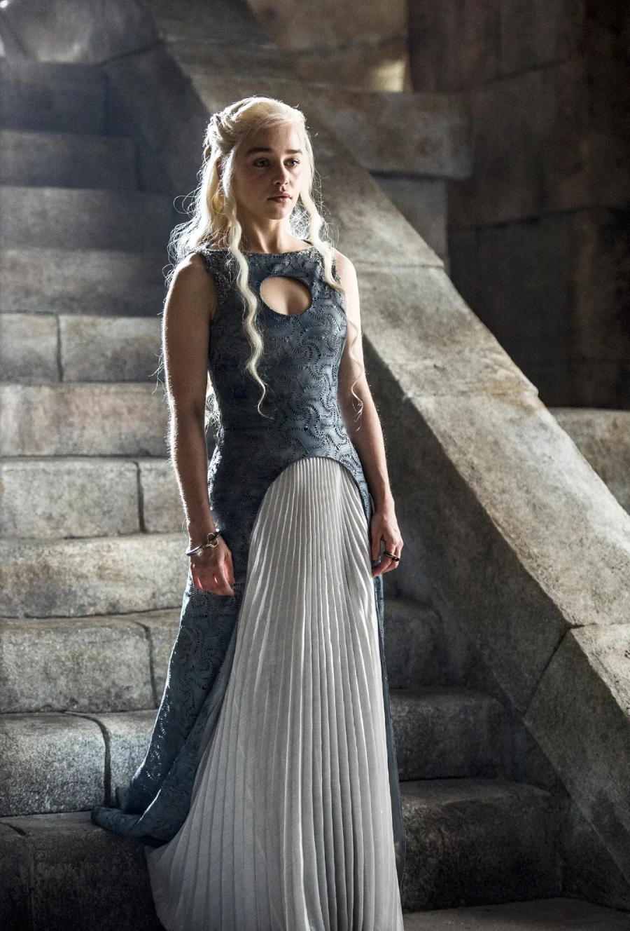 A woman with long, braided blonde hair wearing a gray and white gown stands on stone steps in a dimly lit, medieval-looking setting. She gazes forward with a serious expression.