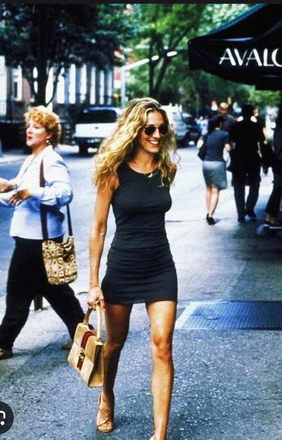 A woman with long blond hair wearing sunglasses and a short black dress walks confidently down a city sidewalk, holding a beige handbag. People and trees line the background, and an “AVALON” awning is visible.