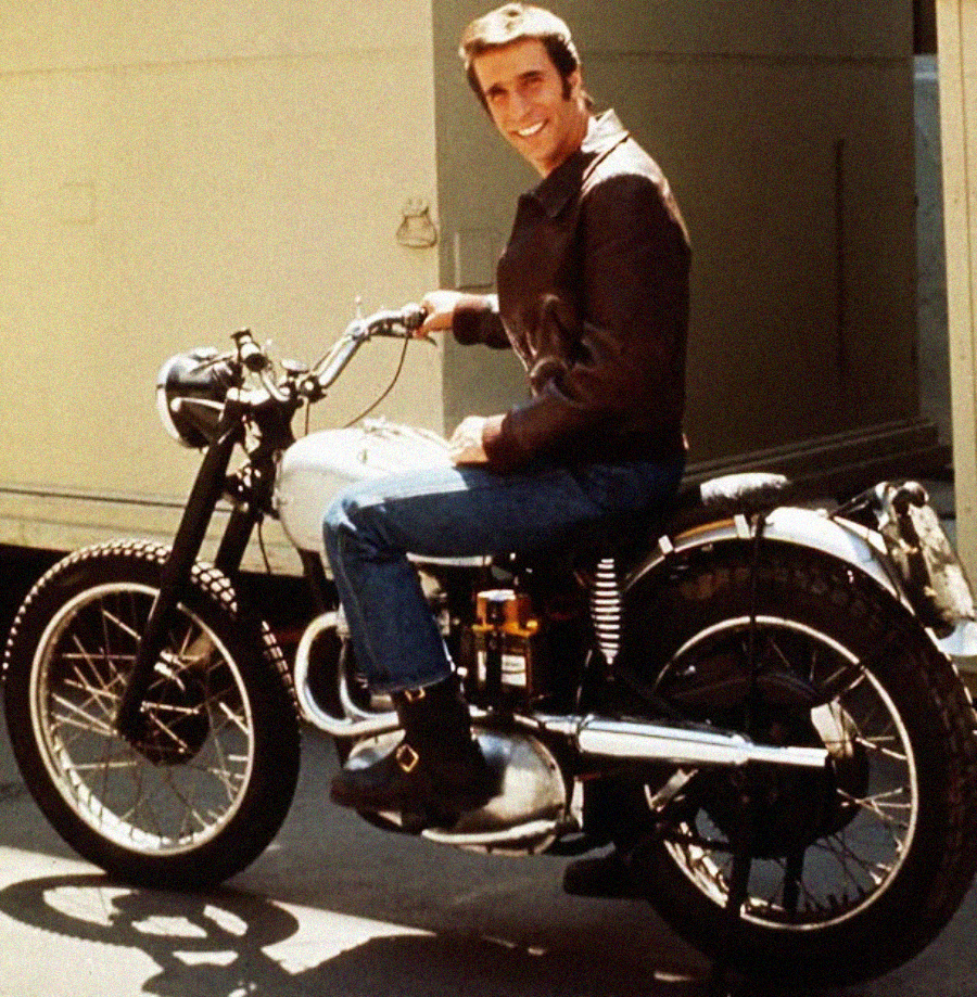 A man wearing a brown leather jacket, blue jeans, and black boots sits smiling on a vintage motorcycle outdoors near a light-colored building.
