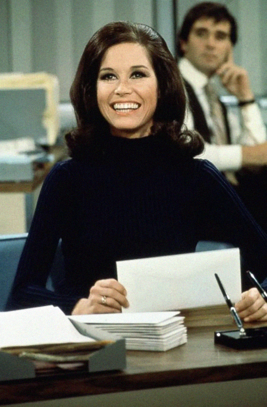 A woman with dark hair in a navy sweater sits at a desk, smiling and holding papers. A man in a shirt and tie sits in the background. The setting appears to be an office, with files, folders, and office supplies on the desk.