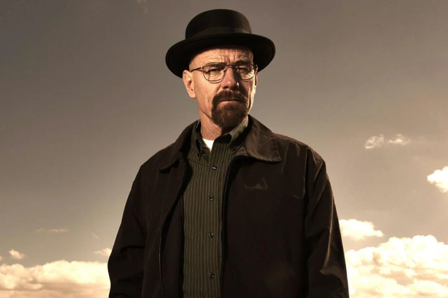 A man with glasses, a goatee, and a black hat stands outside against a cloudy sky, wearing a dark jacket and green shirt, looking seriously at the camera.