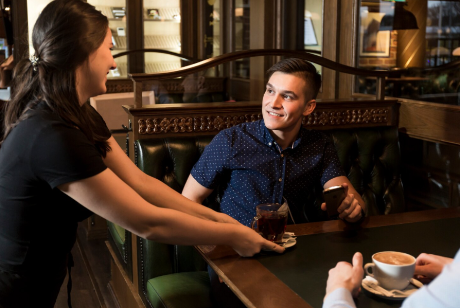 A smiling woman serves drinks to a young man and another person at a cozy café. The man in a blue shirt looks up and smiles, holding his phone, while the table has coffee and tea cups.
