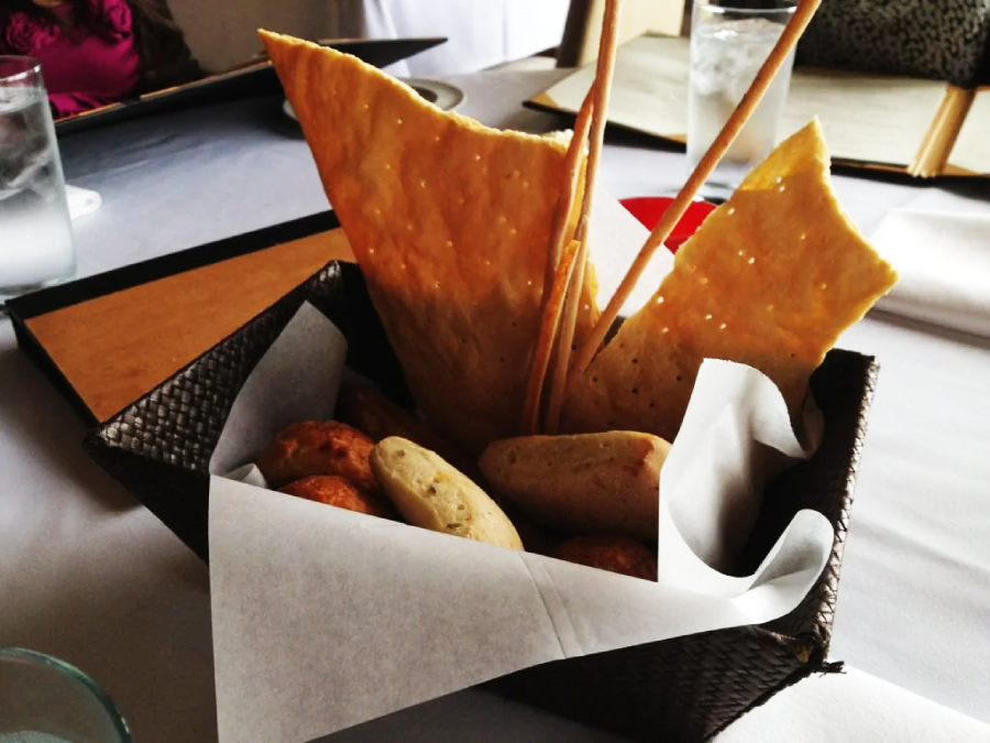 A basket lined with white paper holding assorted bread, including breadsticks, flatbread, and rolls, sits on a restaurant table set with menus and glasses of water.
