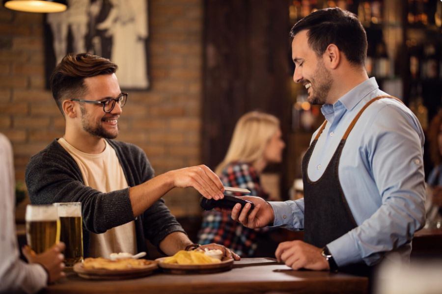 A smiling man pays with his smartphone at a bar while a bartender holds a card reader. There are drinks and food on the table, and other patrons are visible in the background.