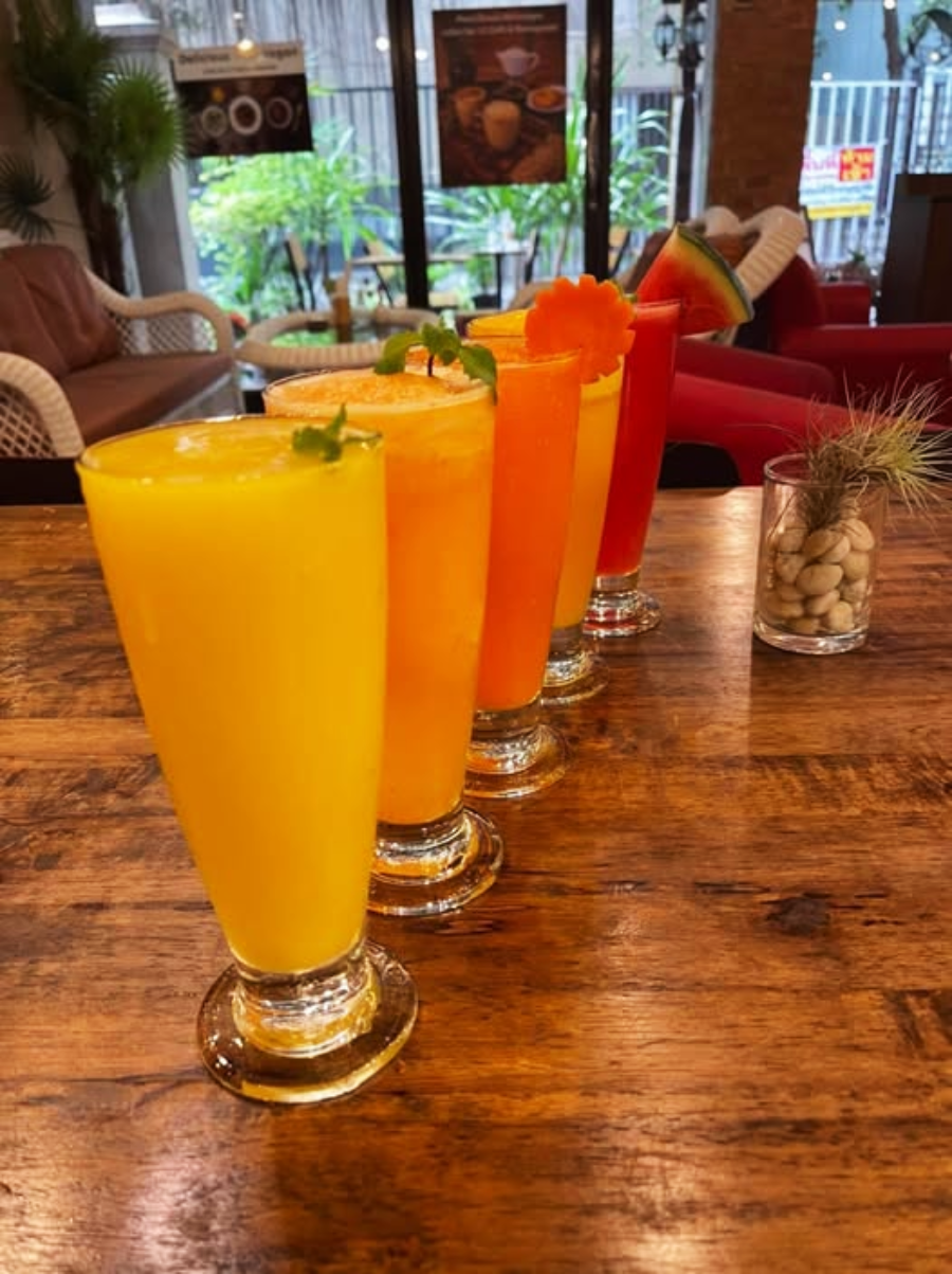 Five colorful fruit smoothies in tall glasses are lined up on a wooden table in a cozy café, each topped with a garnish like mint, a carrot slice, or a watermelon wedge. Greenery and soft chairs are visible in the background.