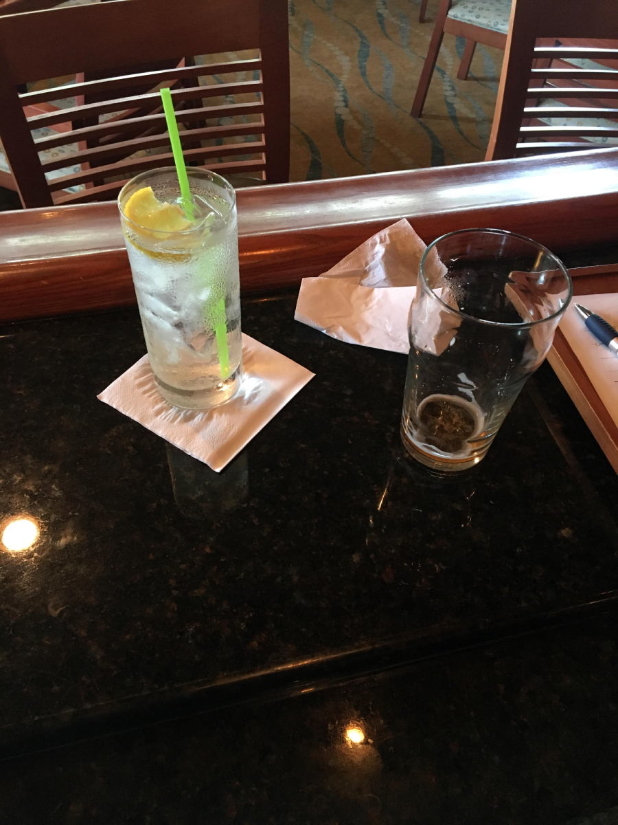 A glass of water with lemon slices and a green straw sits next to an almost empty pint glass on a dark countertop, with napkins and wooden chairs in the background.
