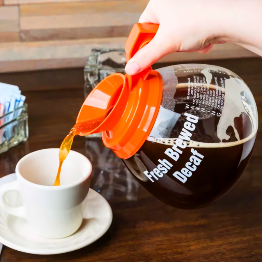 A hand pours coffee from a glass pot labeled "Fresh Brewed Decaf" into a white cup on a saucer, with sugar packets and an ashtray on the wooden table.