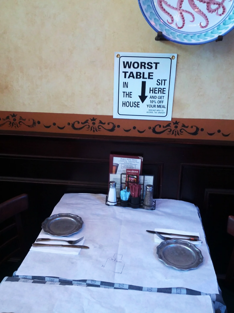 A restaurant table for two with paper placemats, cutlery, and condiments. A sign on the wall above reads, “Worst table in the house. Sit here and get 10% off your meal” with a large arrow pointing down.