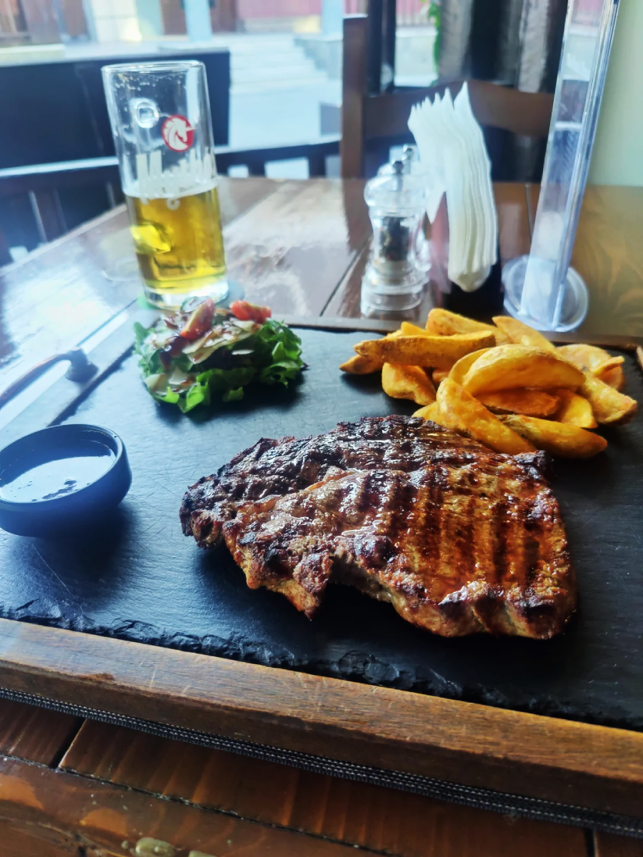 A grilled steak served with thick-cut fries, a small salad, a cup of sauce, and a glass of beer on a wooden table in a restaurant setting.