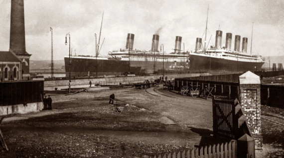 A black-and-white photo shows two large ocean liners with four smokestacks docked side by side at a shipyard, with workers and equipment visible in the foreground.