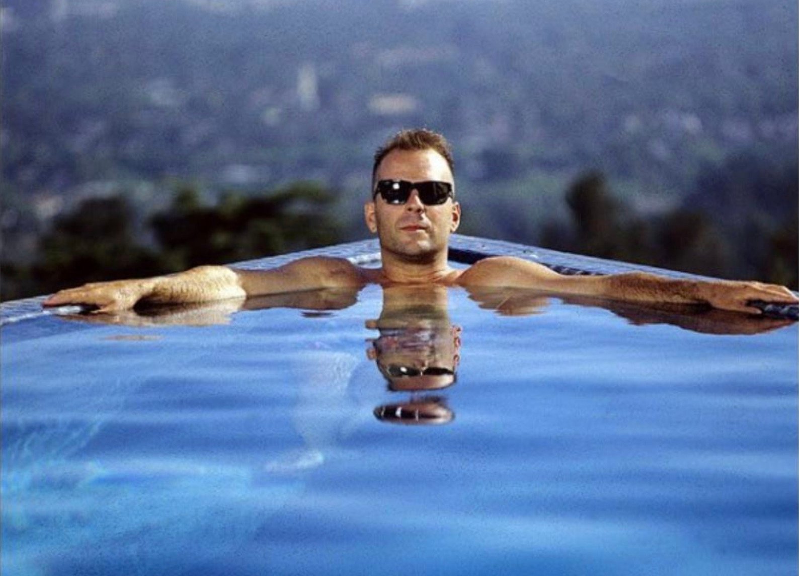 A man wearing sunglasses relaxes with his arms stretched out along the edge of an infinity pool, with a cityscape and greenery blurred in the background. His reflection is visible in the water.