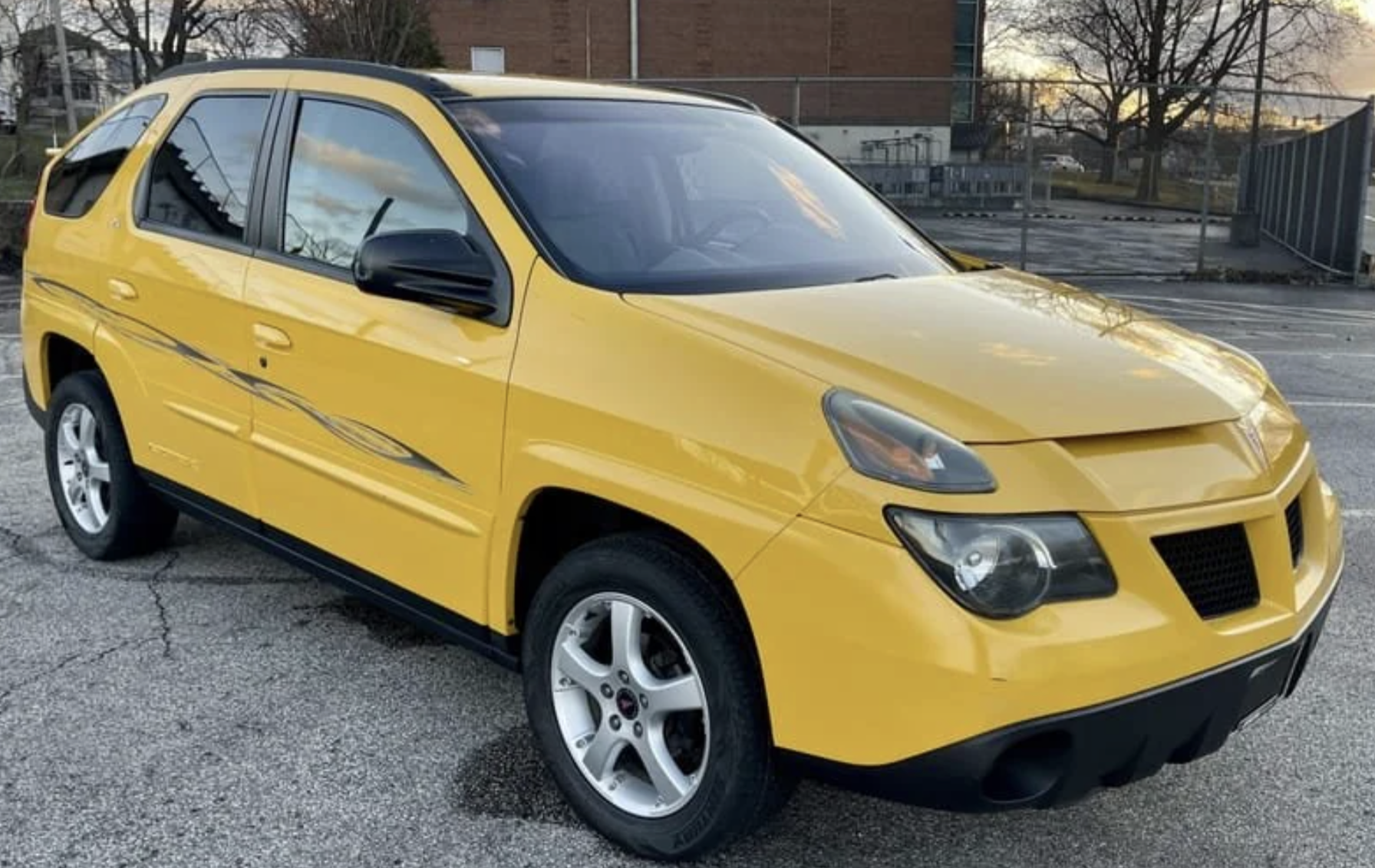 A bright yellow Pontiac Aztek SUV is parked on a paved lot near a chain-link fence, with a brick building and bare trees in the background.