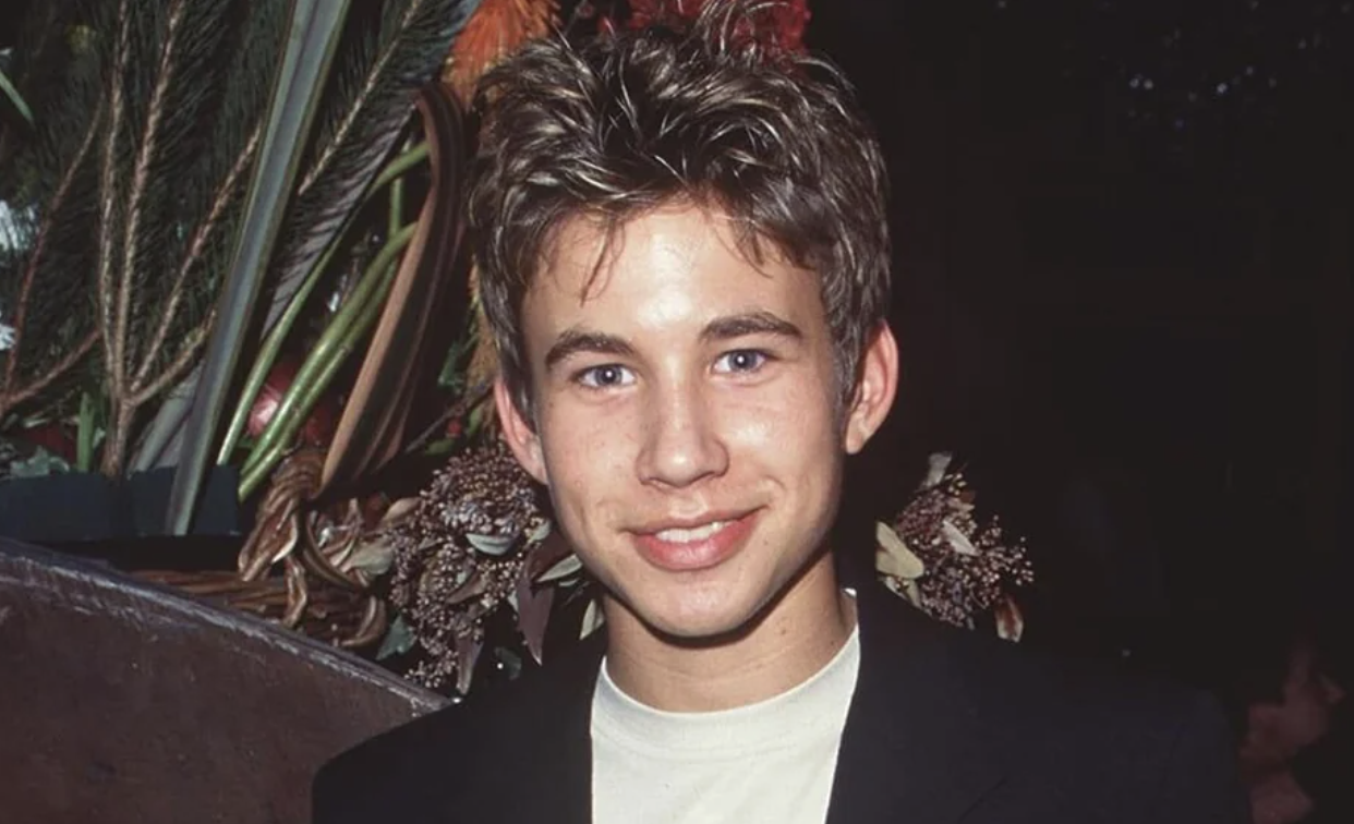 A young man with spiky brown hair smiles at the camera, wearing a light-colored shirt and a dark jacket. Decorative foliage and flowers are visible in the background.