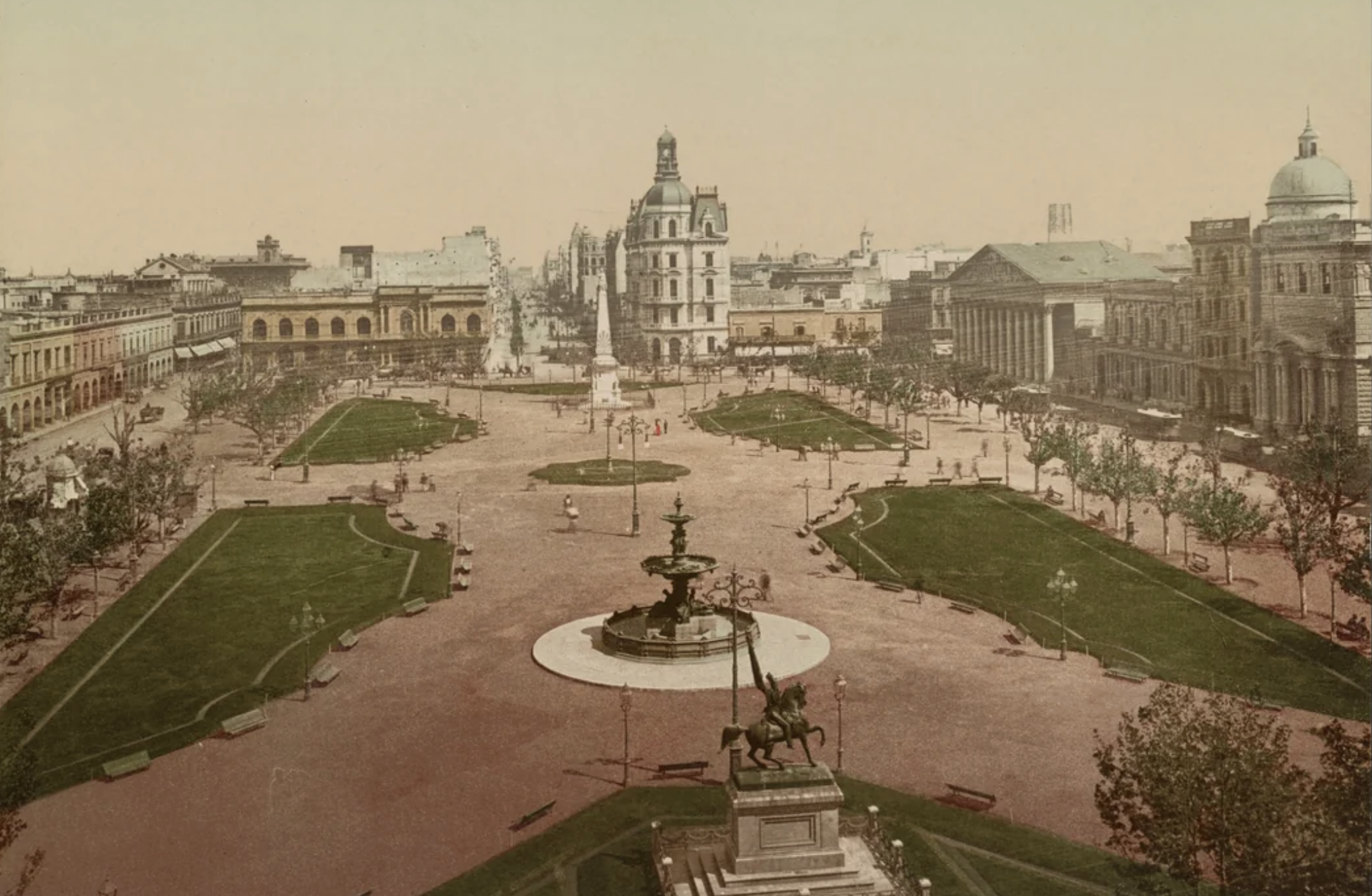 A historic city square with a central fountain, statue, green lawns, and surrounding ornate buildings under a clear sky. Pedestrians are visible walking throughout the plaza.
