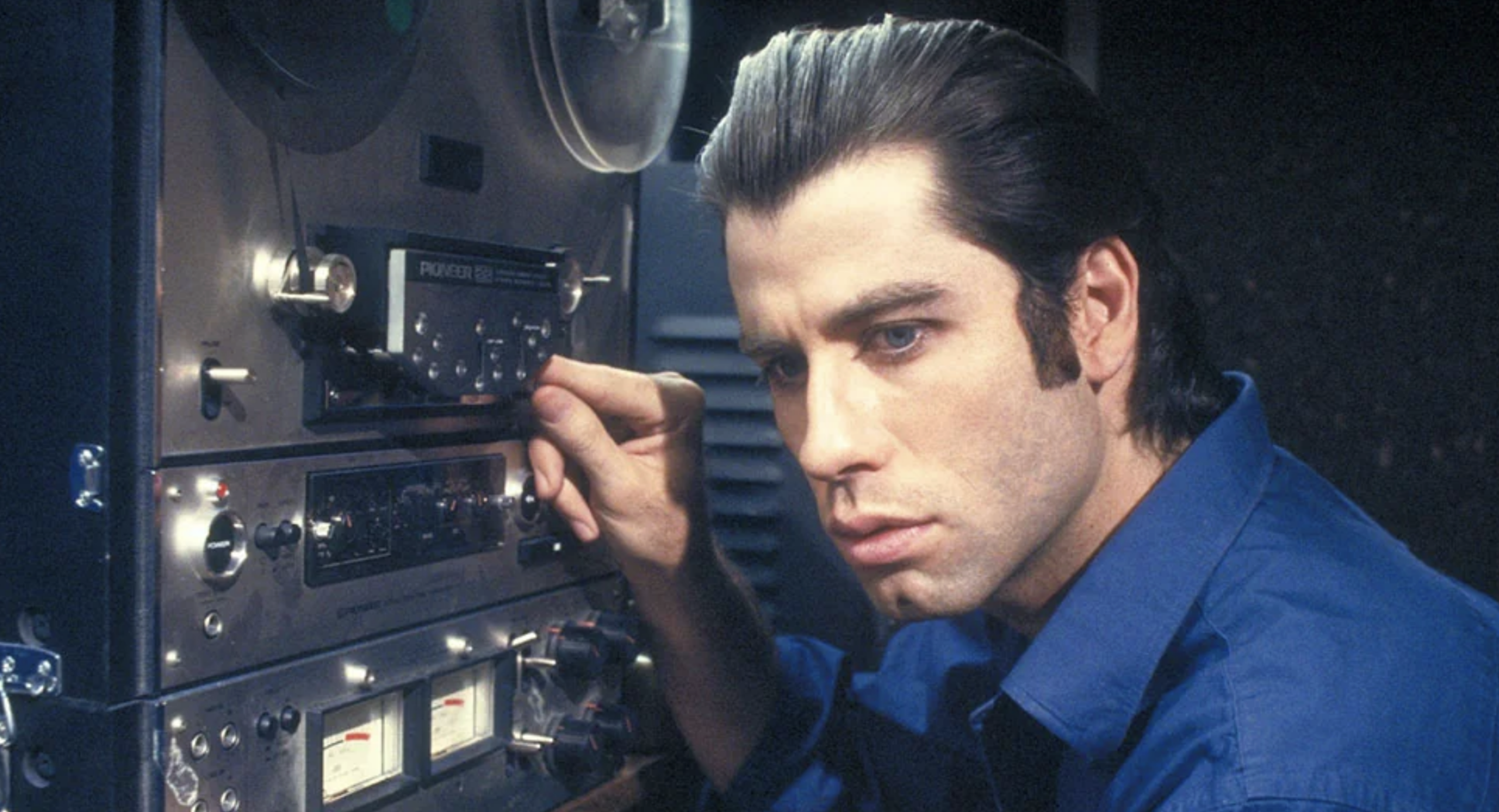 A man with slicked-back dark hair, wearing a blue shirt, operates audio recording equipment, adjusting a dial while focusing intently.