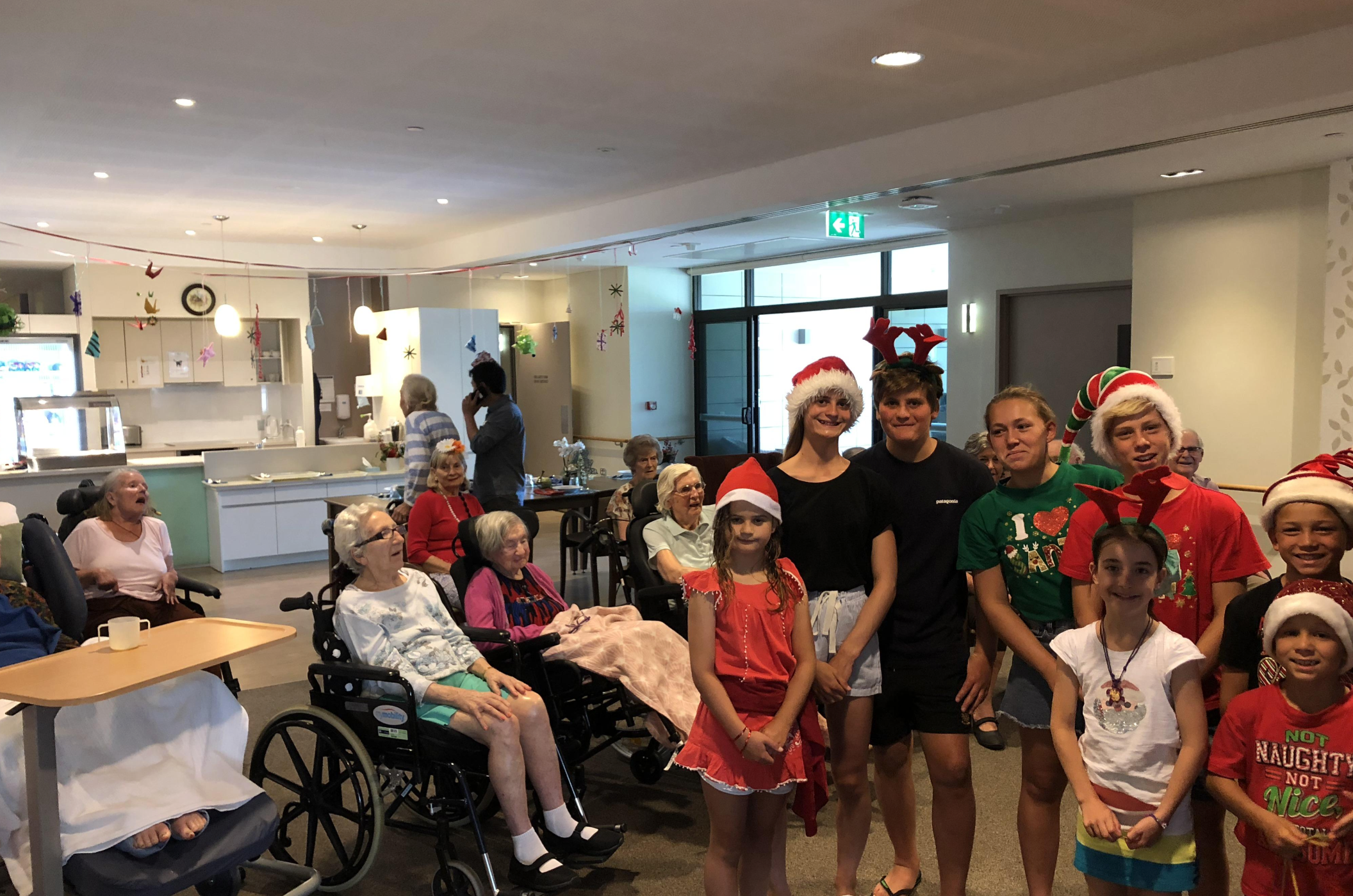 A group of children and adults, some wearing Santa hats, stand and smile in a festive room with Christmas decorations. Several elderly people sit nearby, some in wheelchairs, enjoying the holiday gathering.