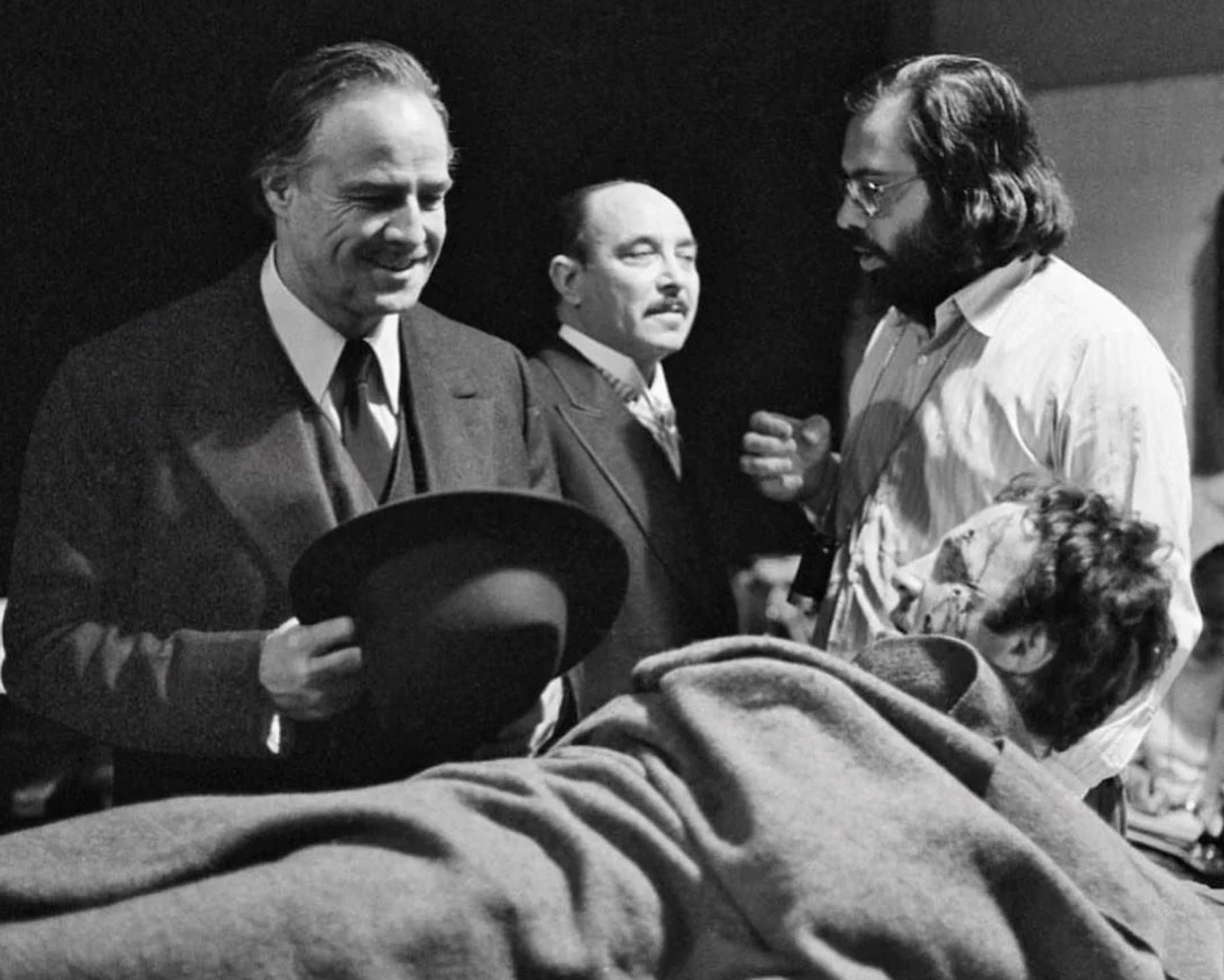 Four men are on a movie set. One man in a suit smiles and holds a hat, another stands behind him, a third man with glasses speaks, and a fourth man lies down covered with a blanket, possibly as part of a film scene.