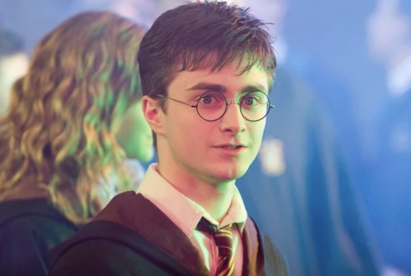 A young person with round glasses, short brown hair, and a striped tie wears a Hogwarts uniform and stands in a softly lit, colorful scene with other students blurred in the background.