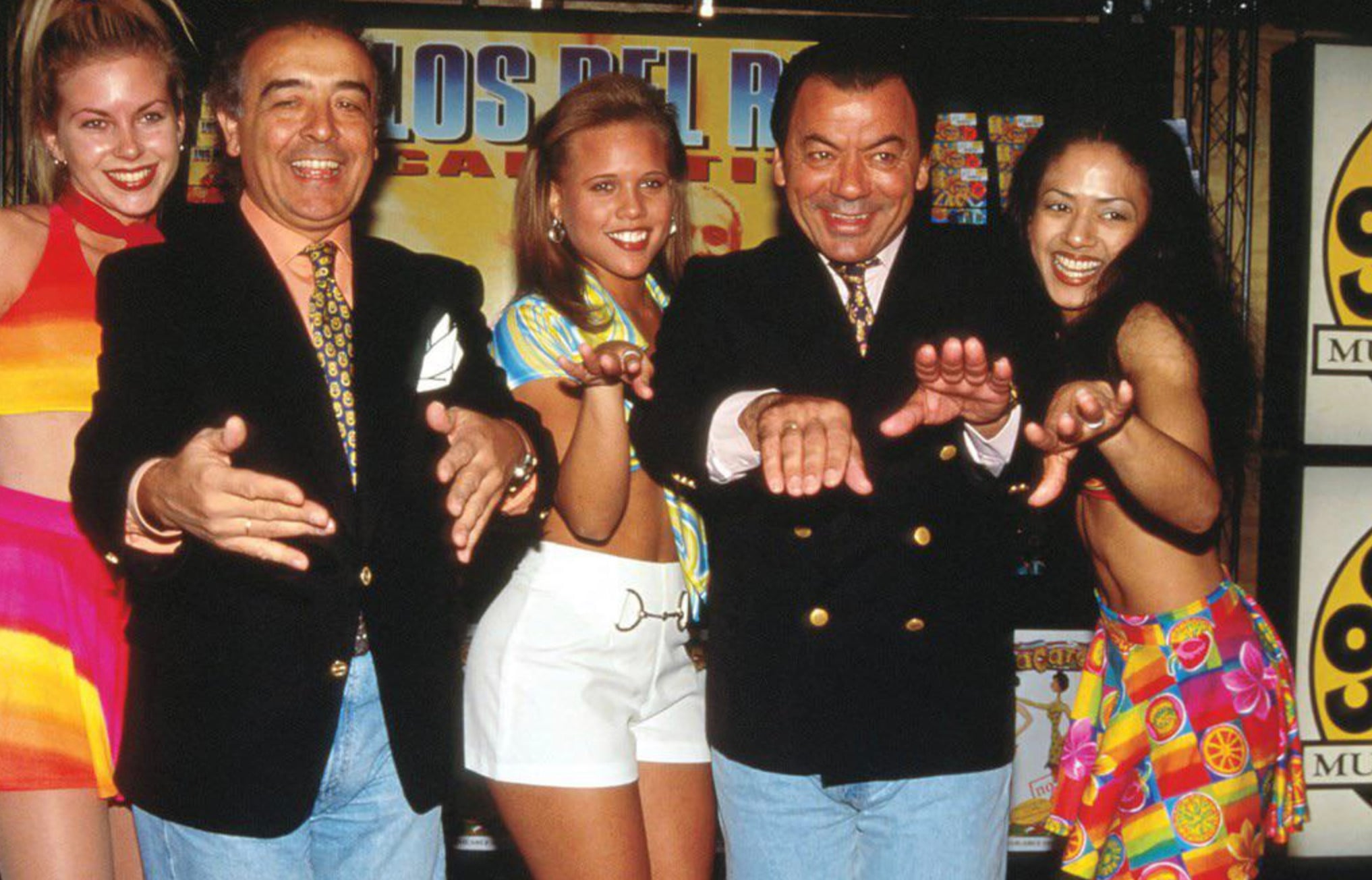 Five people pose energetically in colorful 1990s outfits, smiling and extending their arms towards the camera in front of a bright, festive backdrop with large yellow and orange graphic text.