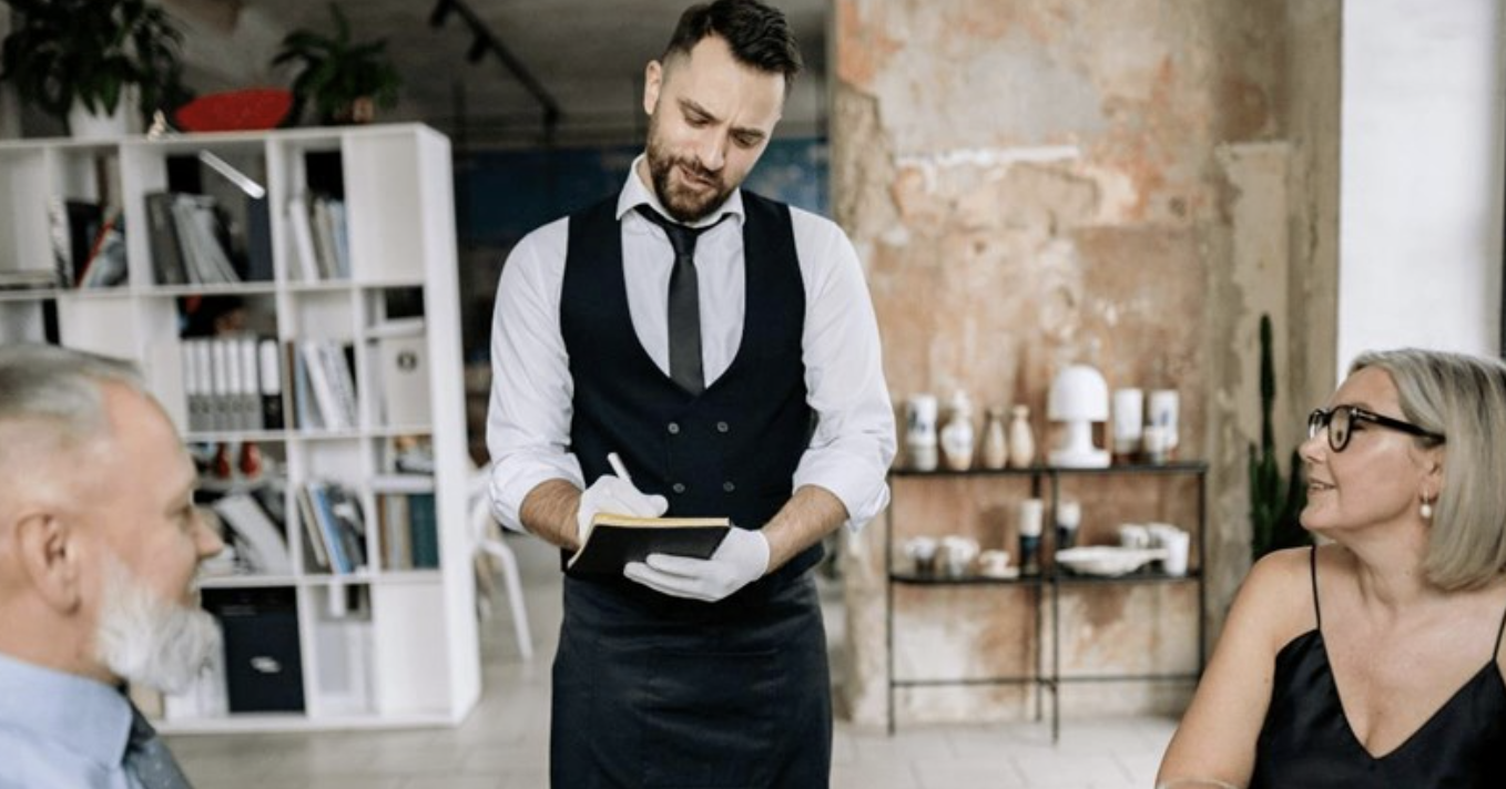 A waiter wearing a vest, tie, and gloves writes on a notepad while taking an order from a seated man and woman in a stylish, rustic restaurant. Shelves and decor are visible in the background.