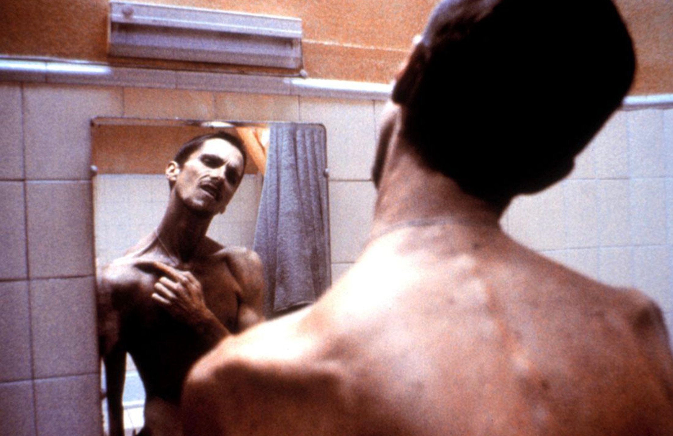 A very thin, shirtless man stands in front of a bathroom mirror, looking at his reflection with a tense expression. The bathroom has tiled walls and a fluorescent light above the mirror.
