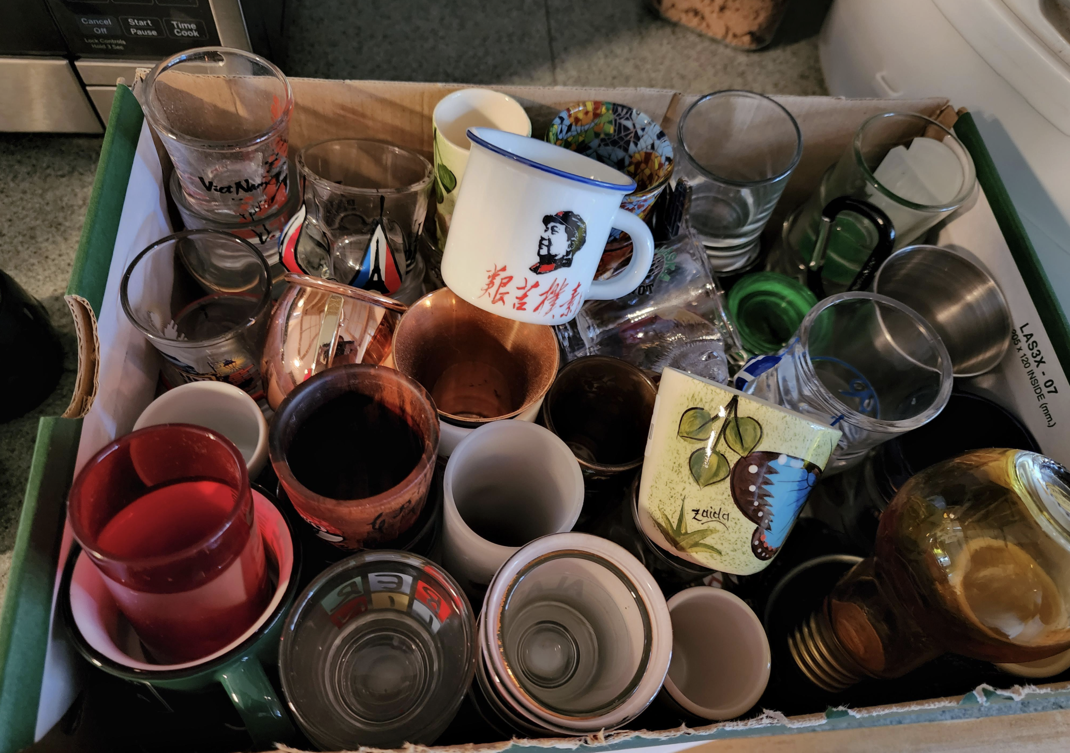 A cardboard box filled with various cups and mugs of different sizes, colors, and designs, including glass, ceramic, and plastic, all packed closely together on a kitchen counter.