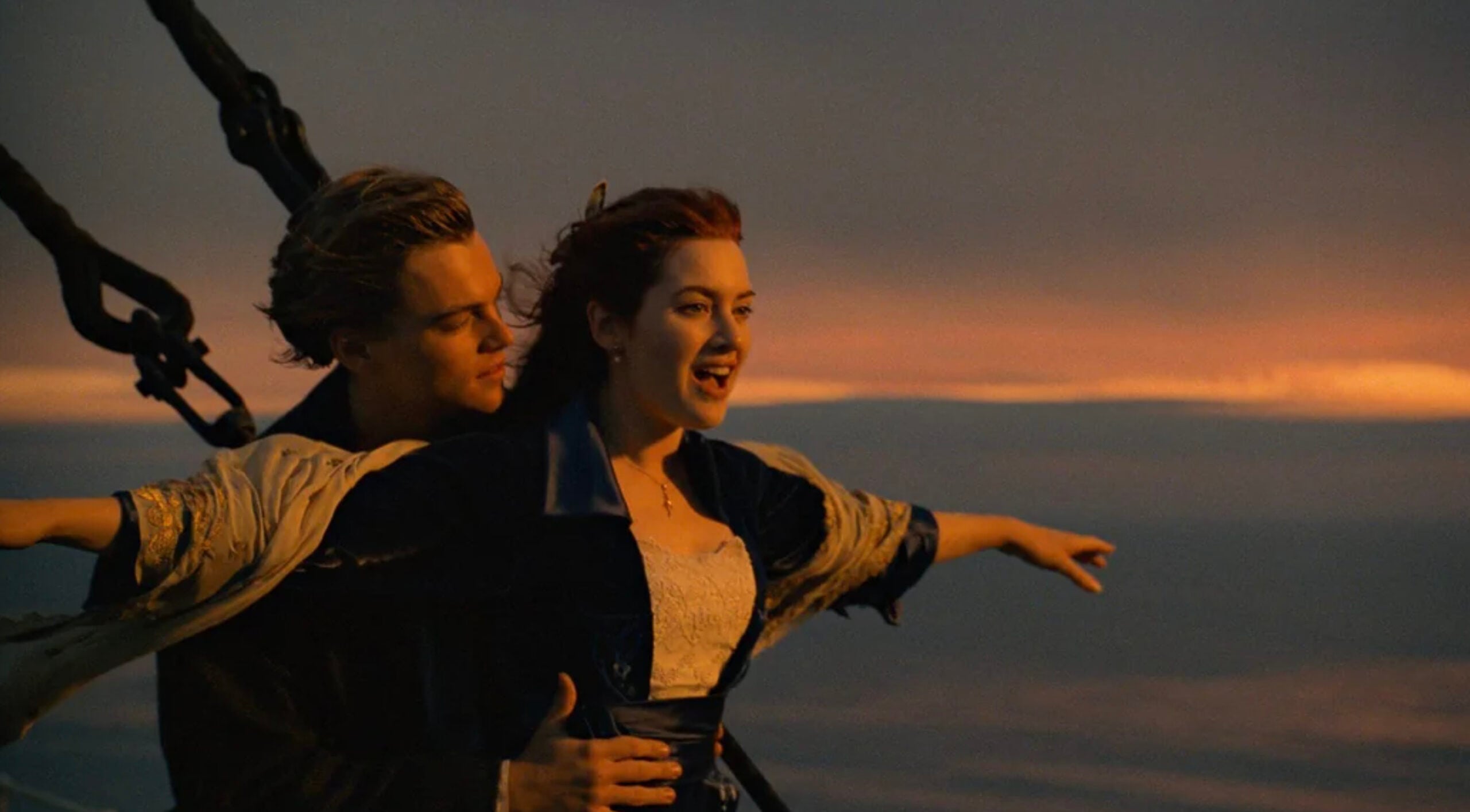 A young man and woman stand at the bow of a ship at sunset. The woman spreads her arms wide while the man stands behind her, holding her waist, as both look joyfully into the distance.