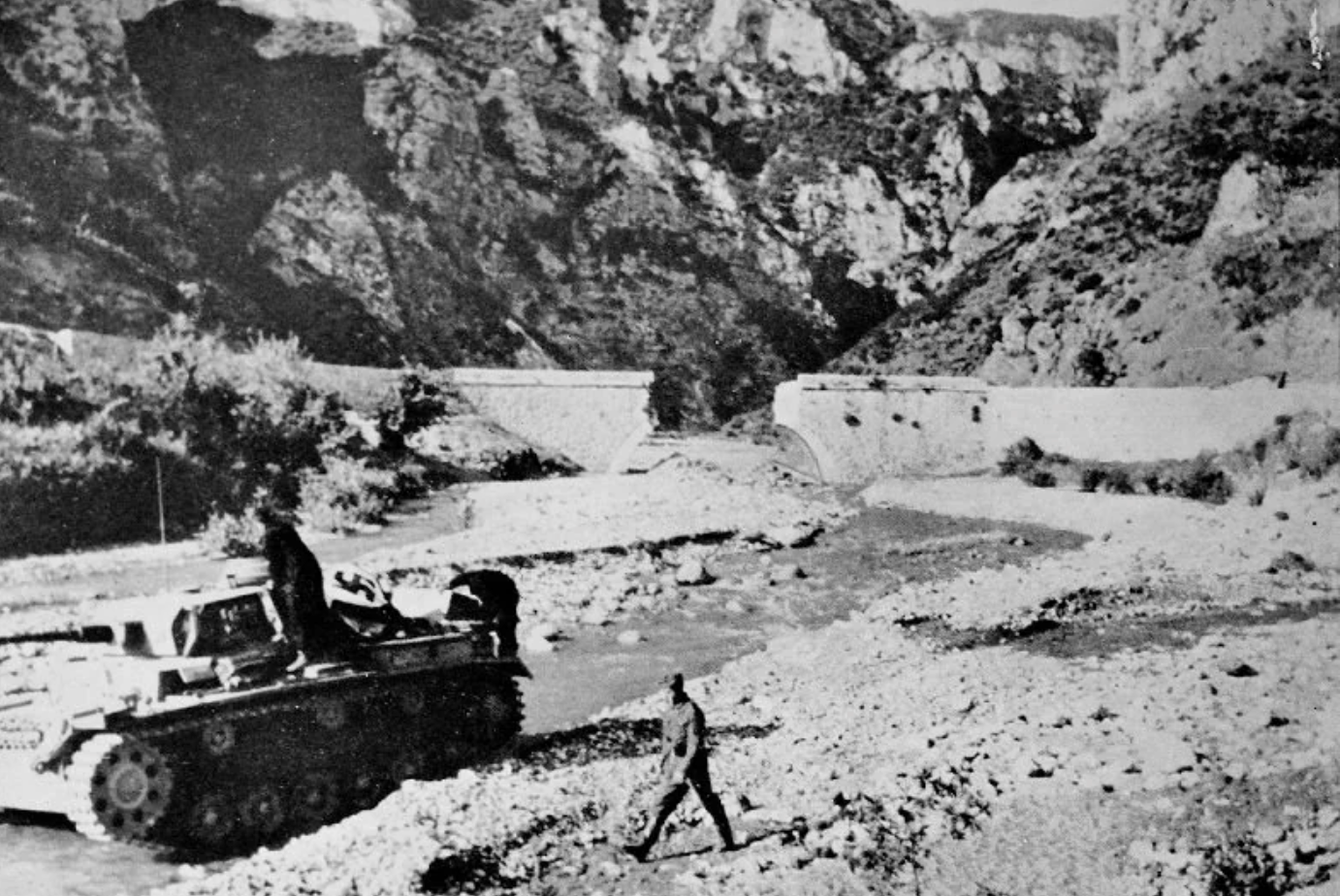A black and white photo shows a tank and two soldiers by a shallow river, with rocky hills and a stone bridge in the background. The setting appears rugged and mountainous.