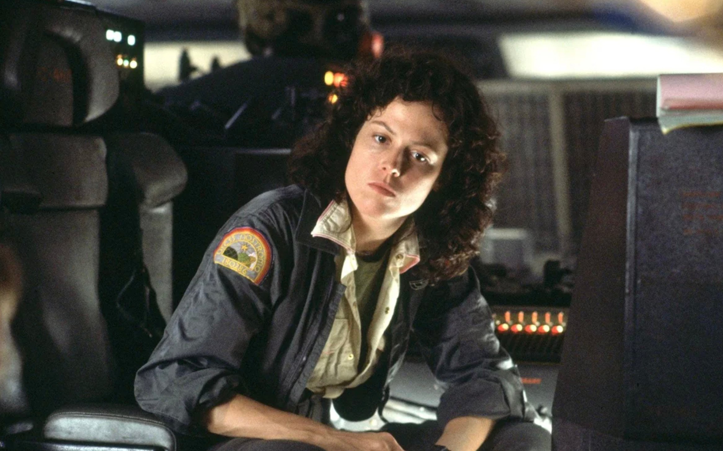 A woman with curly hair in a gray uniform jacket sits in a dimly lit spaceship cockpit, looking intently forward. Control panels with buttons and lights are visible in the background.
