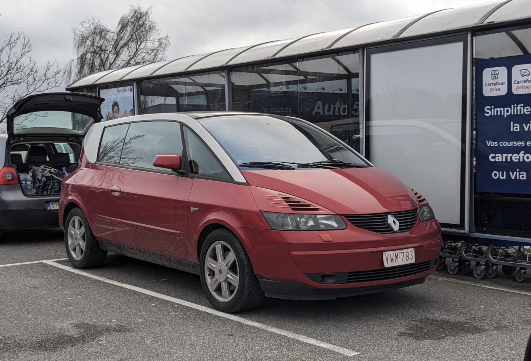 A red Renault Avantime car is parked in a lot near a shopping cart area, with another car next to it and a partially cloudy sky in the background.