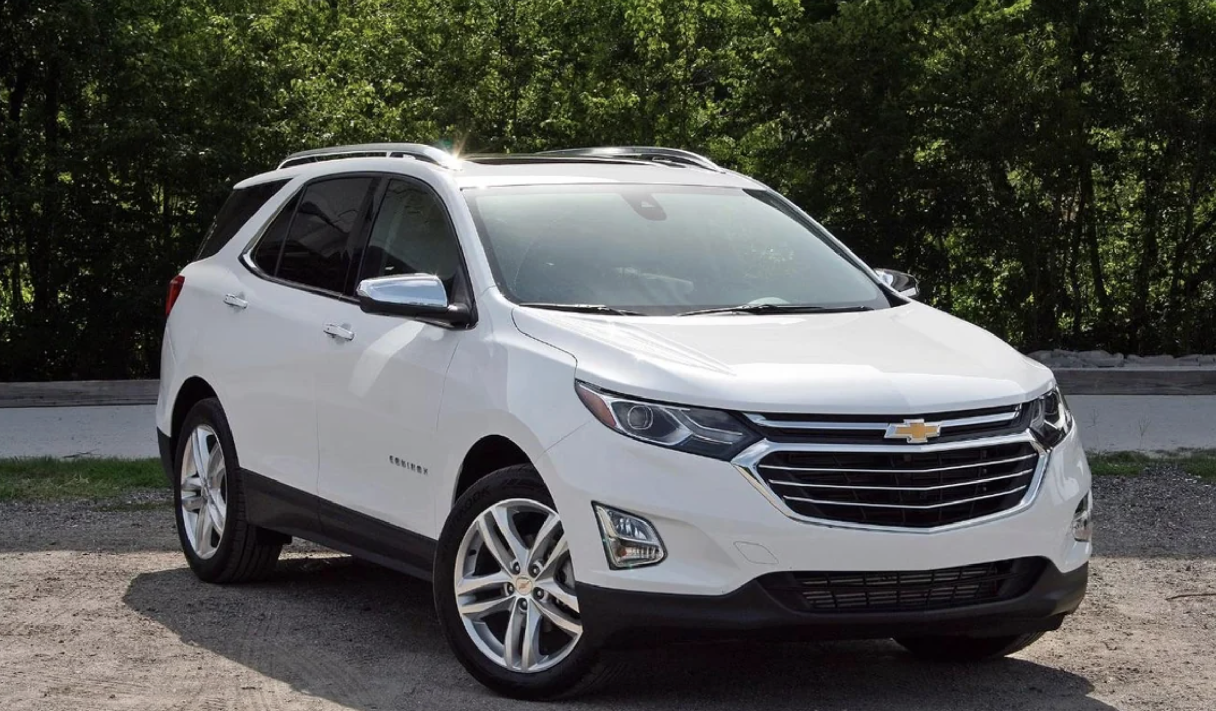 A white Chevrolet Equinox SUV is parked on a gravel surface in front of green trees, under daylight. The vehicle is clean and viewed from the front driver-side angle.