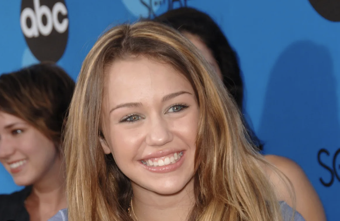 A young woman with long, light brown hair smiles at an event in front of a blue backdrop with ABC logos. She is wearing a light top, and other people are visible in the background.