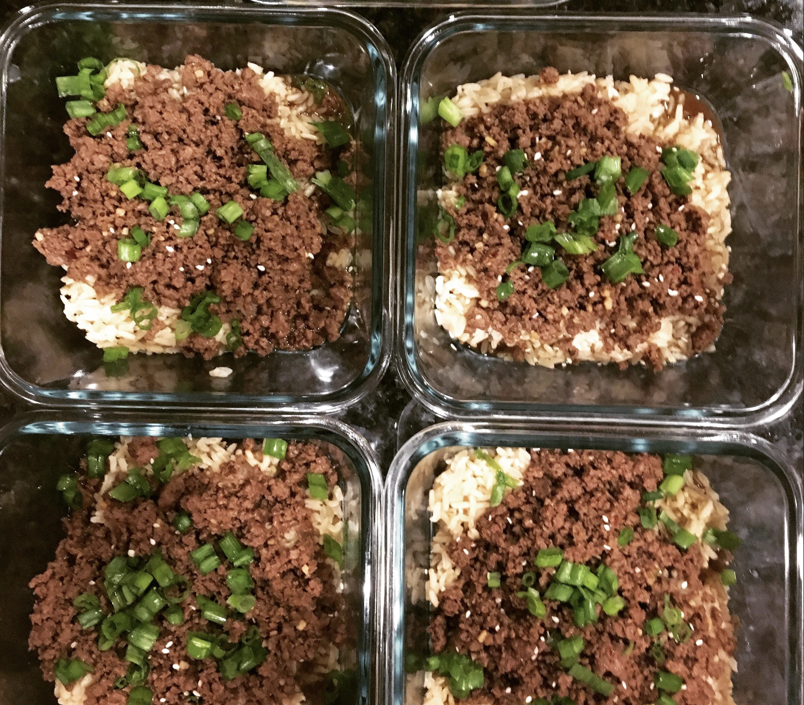Four glass meal prep containers filled with rice, topped with cooked ground beef, chopped green onions, and a sprinkle of sesame seeds. The containers are arranged in a two-by-two grid.