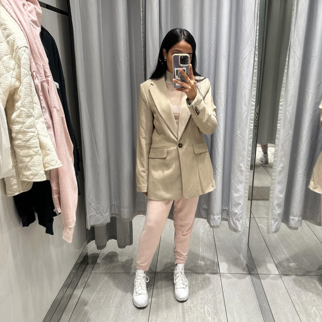 A woman stands in a fitting room taking a mirror selfie. She is wearing a beige blazer over a light pink outfit and white sneakers. Clothing items hang on the wall to her left, and grey curtains are behind her.