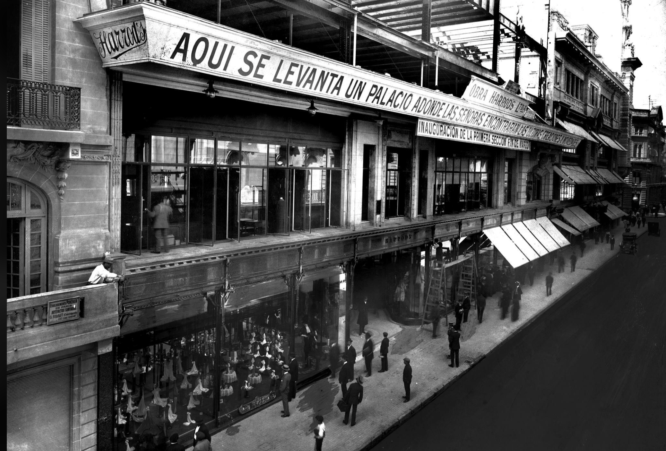 Black and white photo of a city street with people walking by a large building under construction. A sign in Spanish reads, "AQUI SE LEVANTA UN PALACIO," indicating a new palace or grand building is being erected.