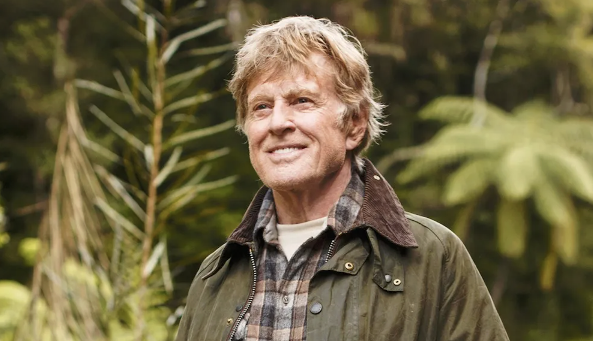 An older man with tousled light hair smiles outdoors, wearing a green jacket over a plaid shirt. Lush green trees and plants create a forested background.