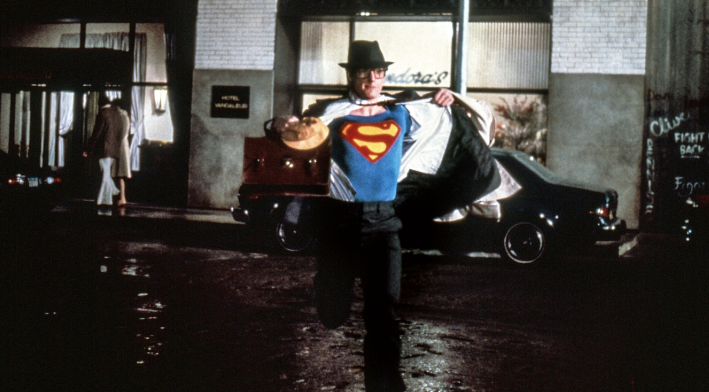 A man in a suit and hat runs down a city street at night, pulling open his shirt to reveal a Superman costume underneath.