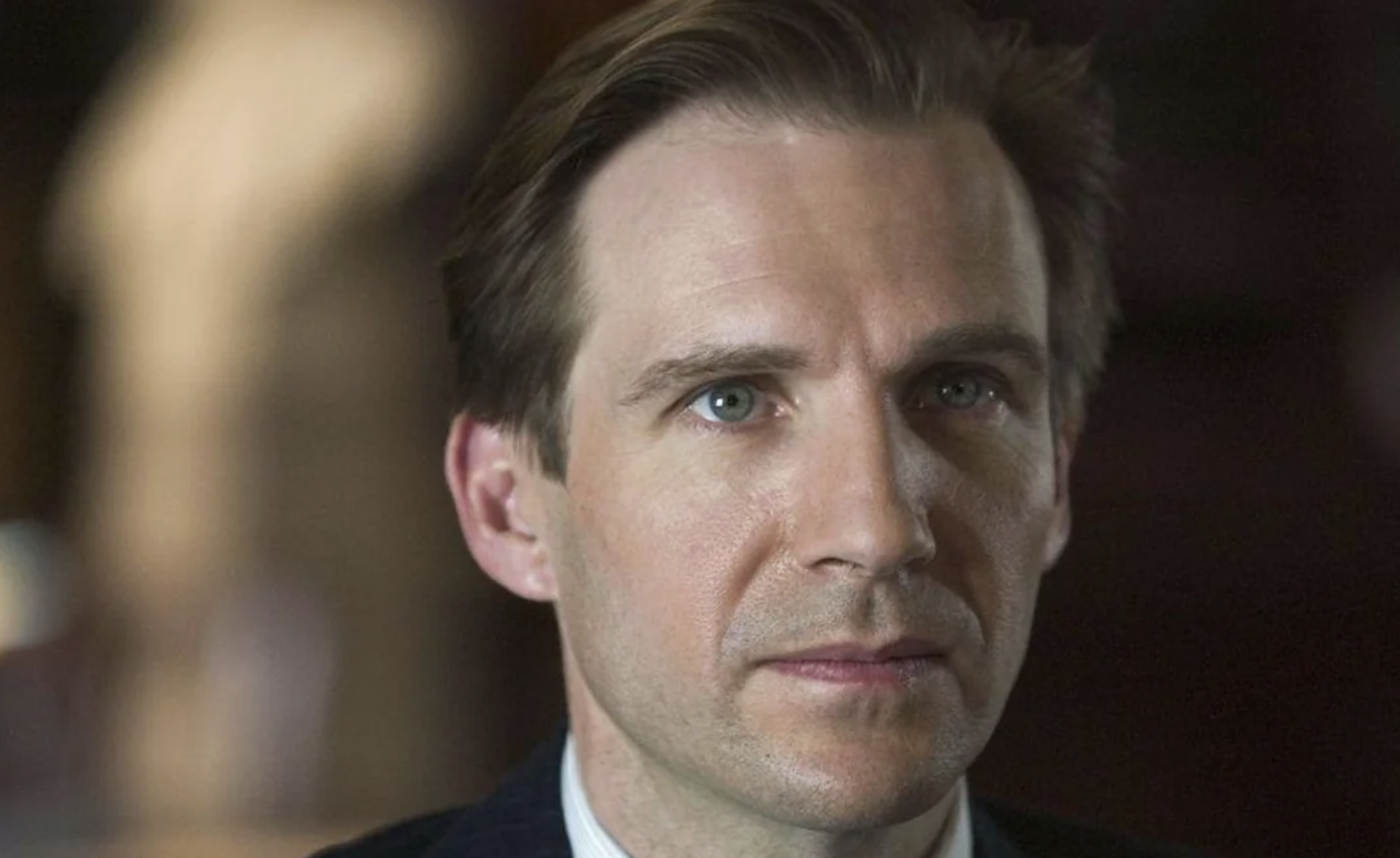 A man with short brown hair and blue eyes looks directly at the camera with a neutral expression. He is wearing a dark suit and a light-colored shirt. The background is softly blurred.