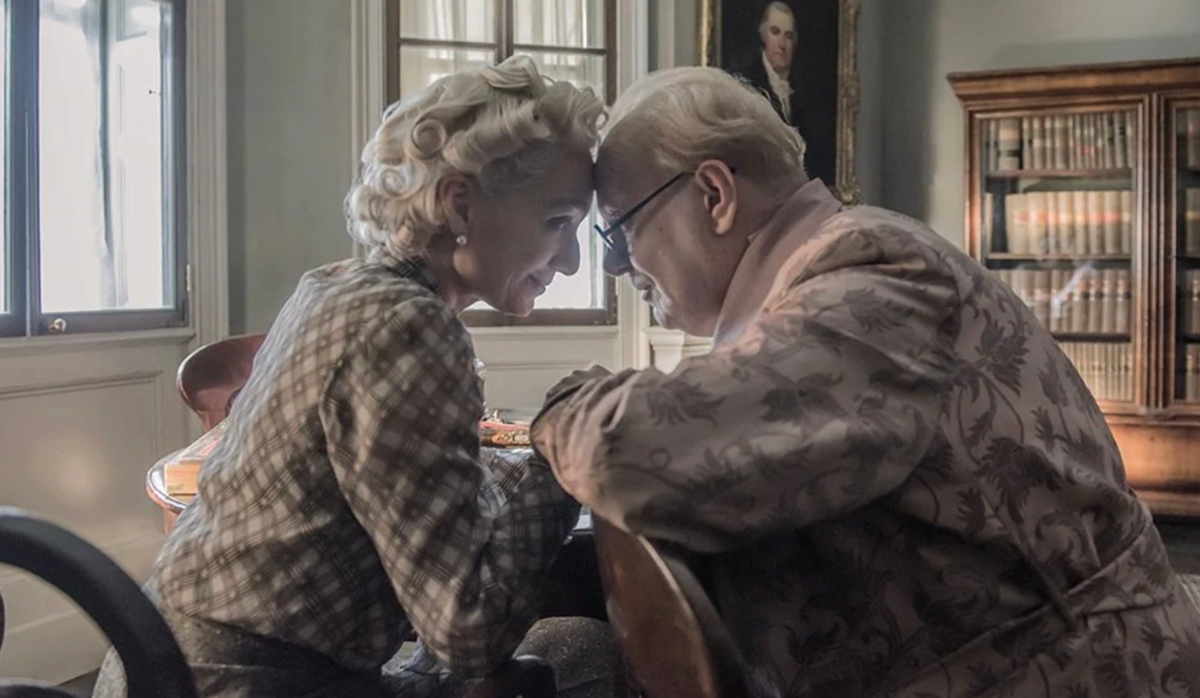 An elderly couple sits close together, foreheads touching and smiling warmly in a cozy room with bookshelves, a large window, and a portrait in the background.