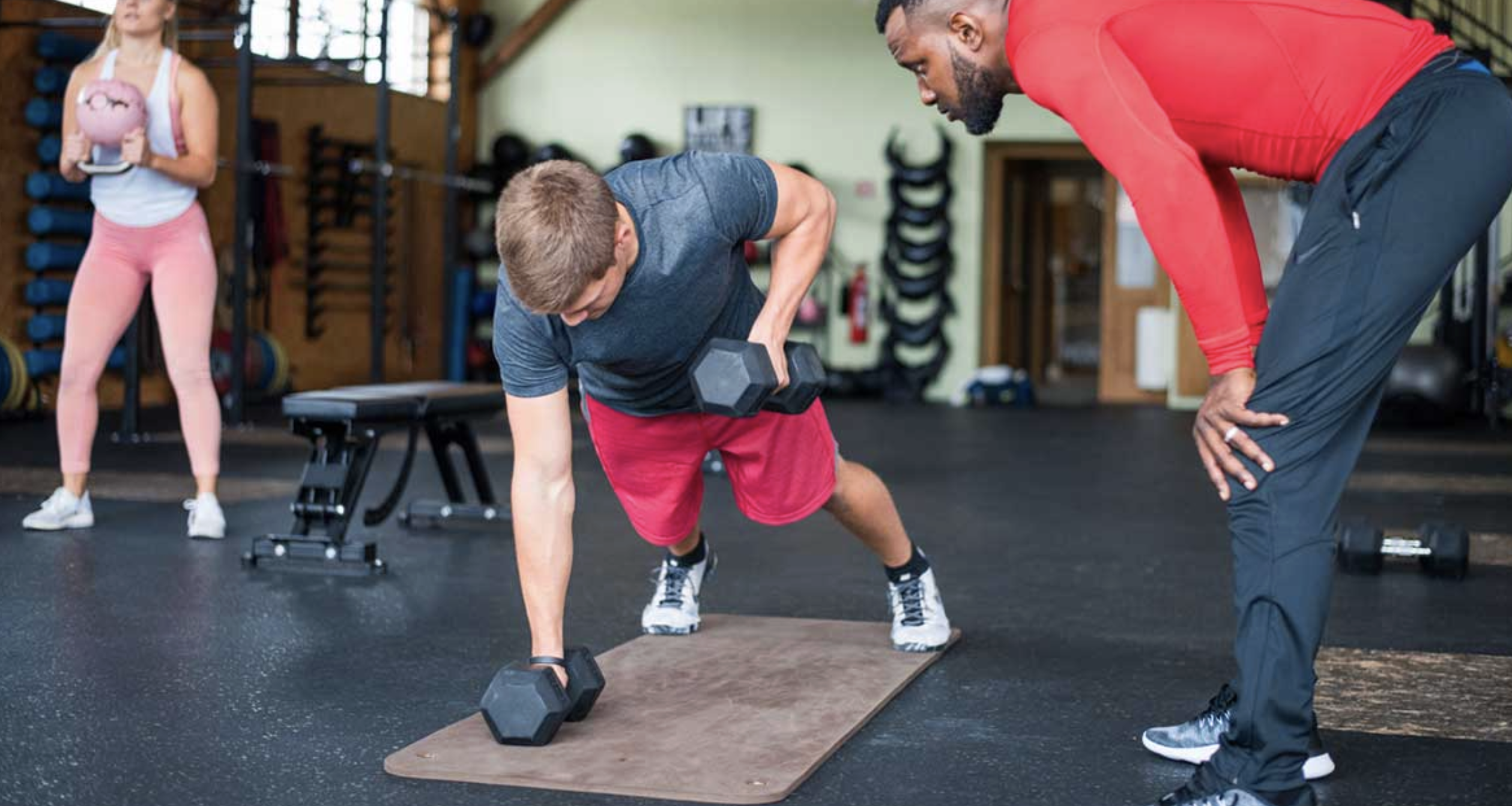 A man in athletic wear performs a renegade row with dumbbells on a mat as a trainer supervises him in a gym. A woman holding a medicine ball stands in the background. Fitness equipment is visible throughout the gym.