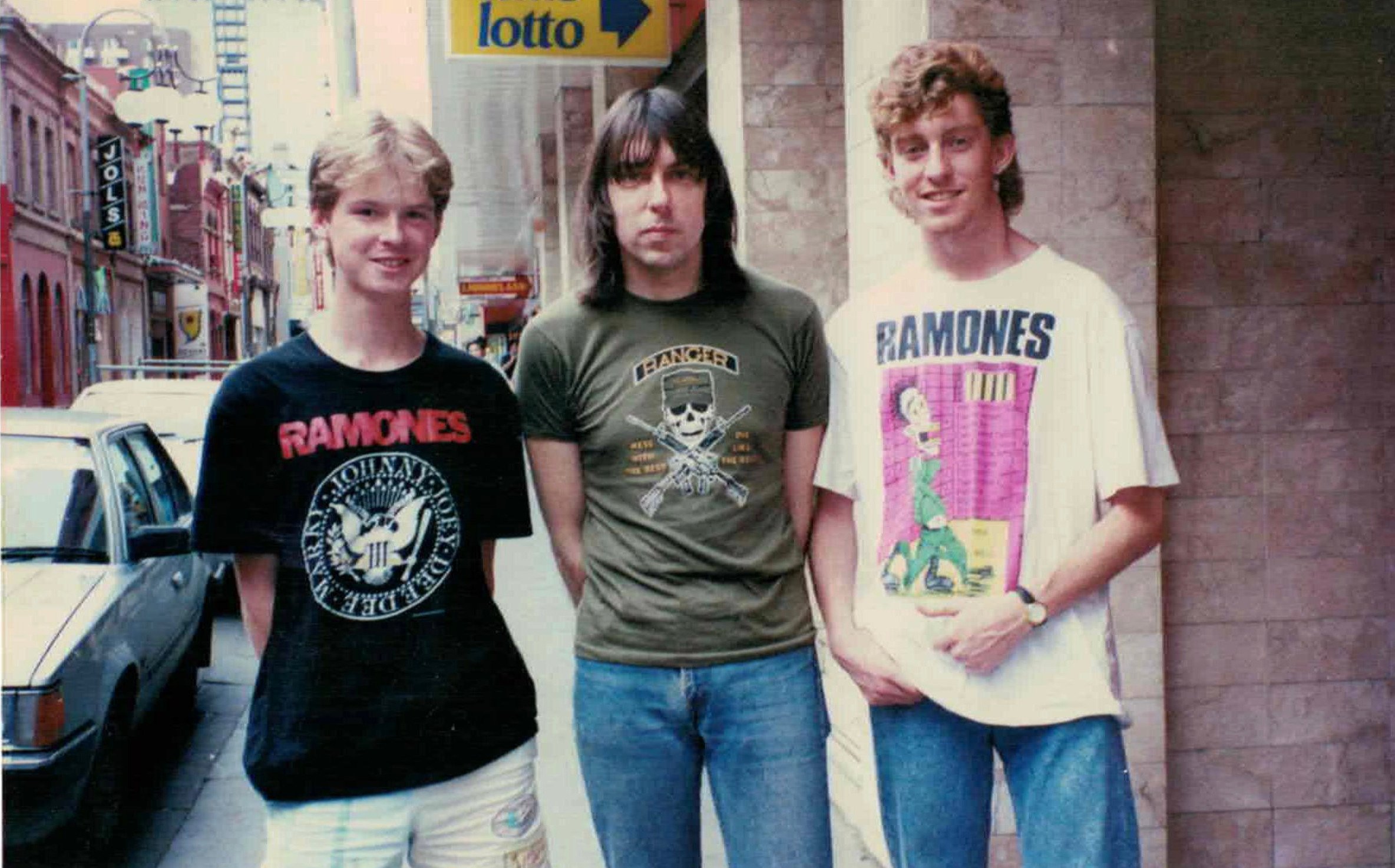 Three young men stand on a city sidewalk near a wall; two wear Ramones t-shirts, and the third wears a green shirt with skull graphic. A car is parked on the street, and there is a "lotto" sign above them.