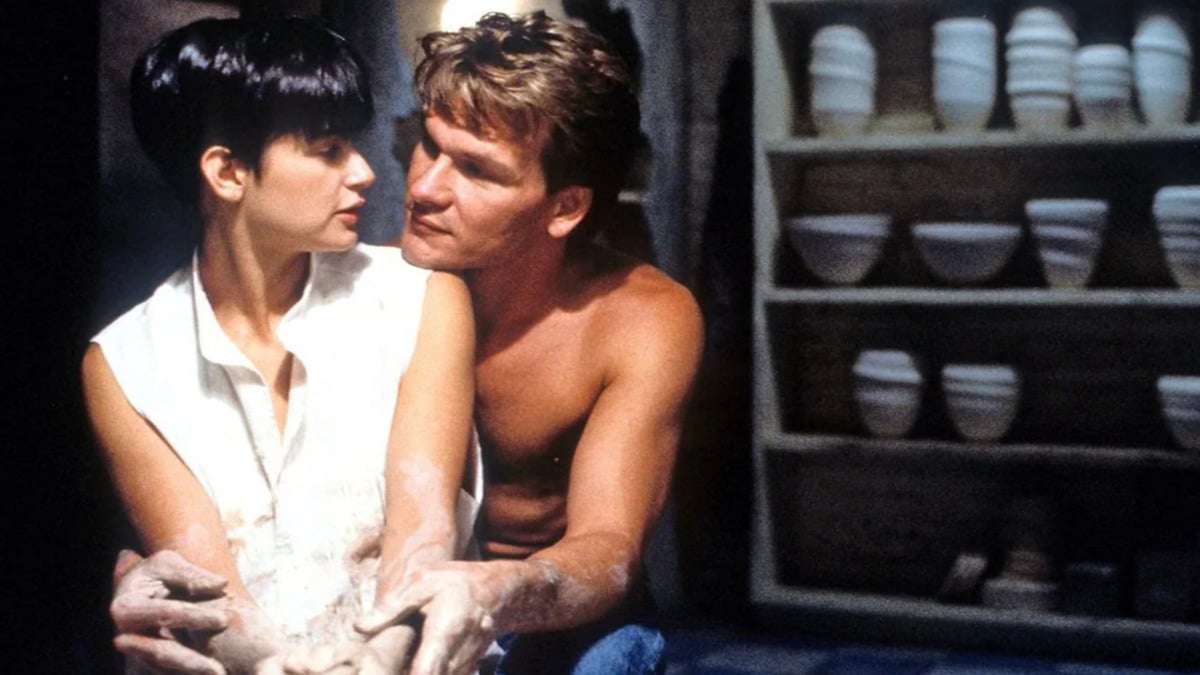 A woman with short dark hair shapes clay on a pottery wheel as a shirtless man sits behind her, guiding her hands. They gaze at each other closely. Several pottery pieces are on shelves in the background.