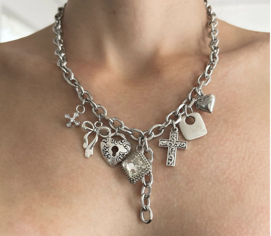 A silver chain necklace with several charms, including a heart, a cross, a flower, a lock, a key, lips, a rectangle, and a crown, worn around a person's neck against bare skin.