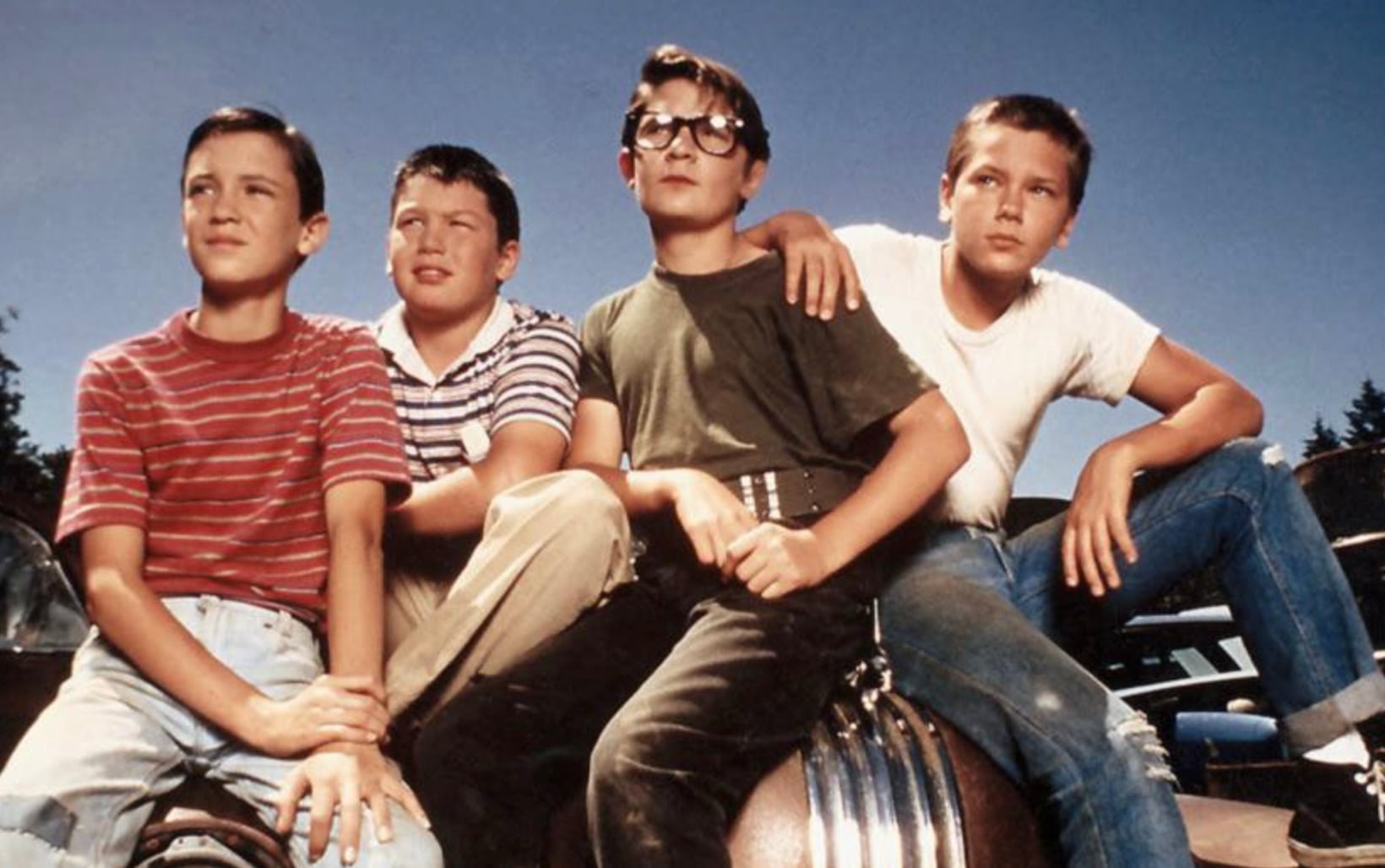 Four boys sit close together outdoors on a sunny day, looking into the distance. They wear casual 1950s-style clothes: striped and plain shirts, jeans, and glasses. They appear thoughtful and united as friends.
