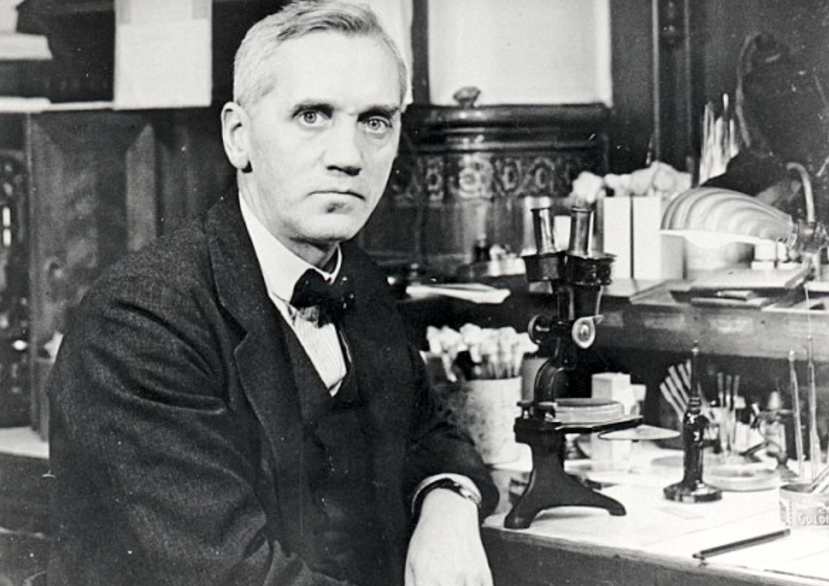 A man in a suit sits at a laboratory desk beside a microscope and various scientific equipment, looking toward the camera in a vintage black-and-white photo.