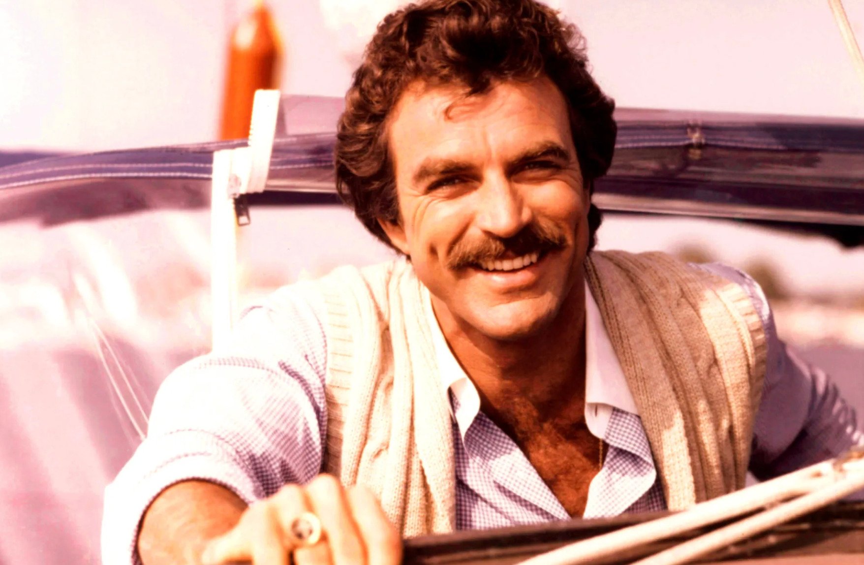 A smiling man with curly brown hair and a thick mustache sits in a convertible car, wearing a light sweater over a blue shirt. The background is bright and sunlit, suggesting a warm, carefree day.
