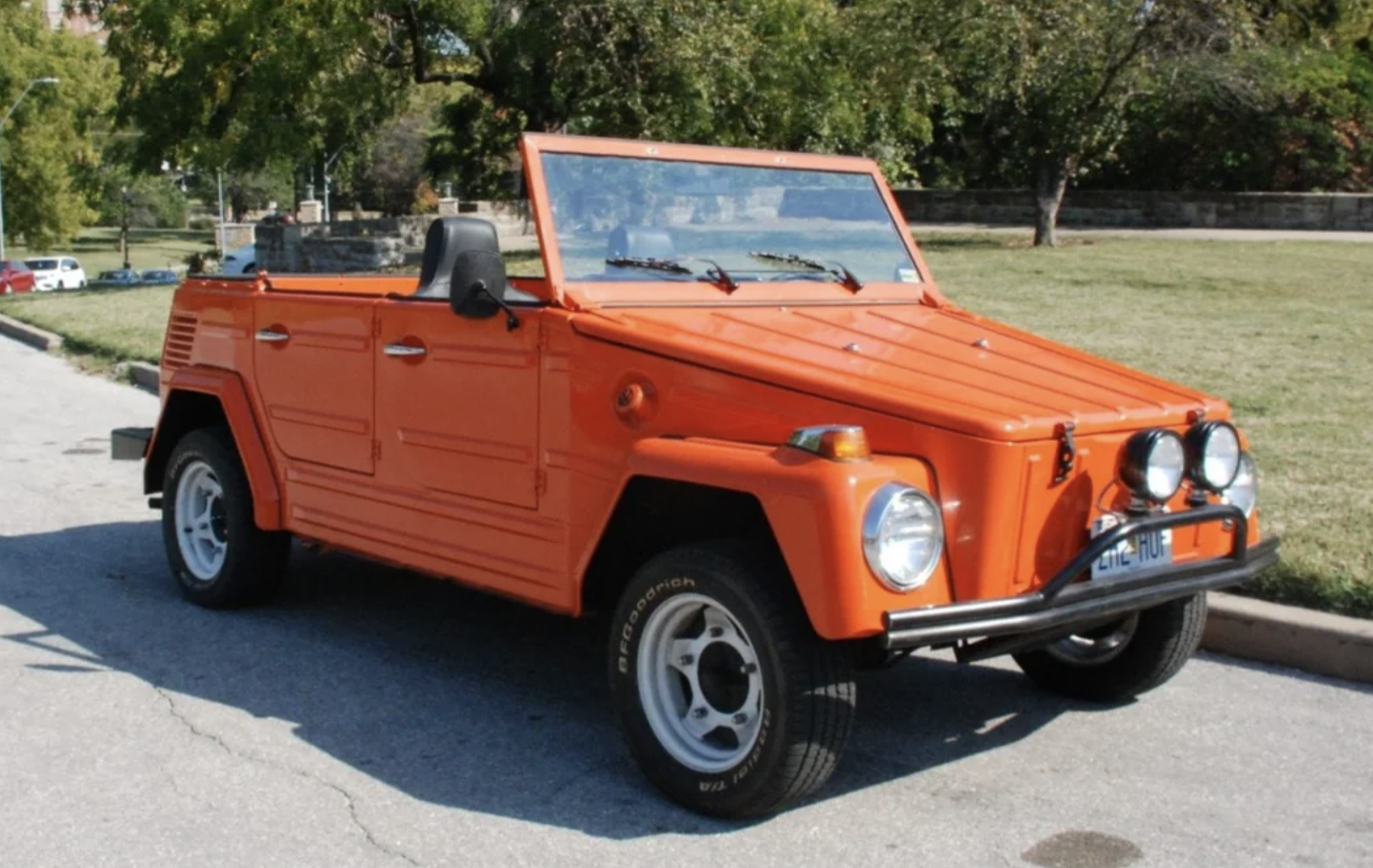 A bright orange vintage convertible car is parked on a street near a grassy area with trees in the background. The car has a boxy design and round headlights.