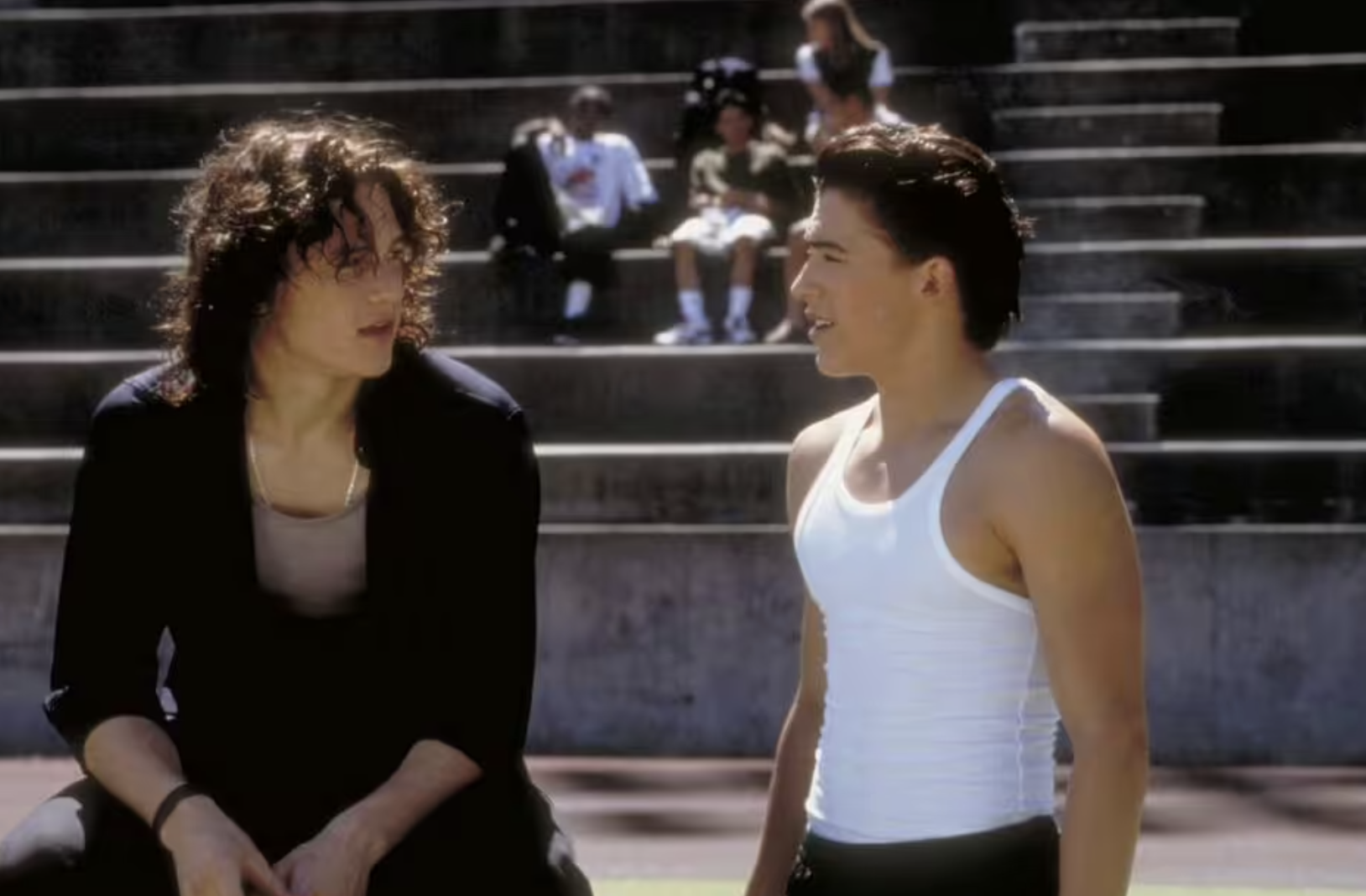 Two young men talk on outdoor bleachers; one has long curly hair and wears dark clothes, the other has short hair and wears a white tank top. Several people sit in the background on higher bleacher rows.
